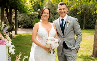 A bride in a white gown and a groom in a gray suit smiling outdoors in a garden setting.