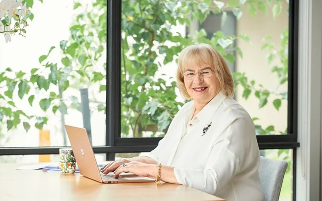 Woman smiling at a table with a laptop, in a room with large windows and greenery outside.