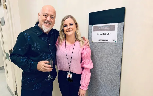 Man holding a glass and woman in a pink shirt posing near a sign reading "Bill Bailey."