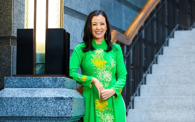 A woman in a green traditional dress standing by stairs, smiling, with a modern building background.