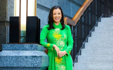 A woman in a green traditional dress standing by stairs, smiling, with a modern building background.