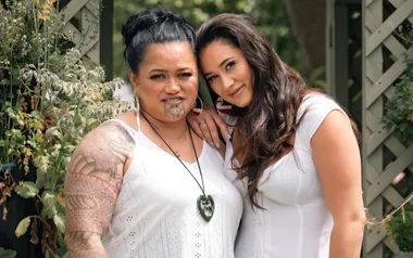 Two women in white dresses stand closely together, smiling in a garden with greenery around them.