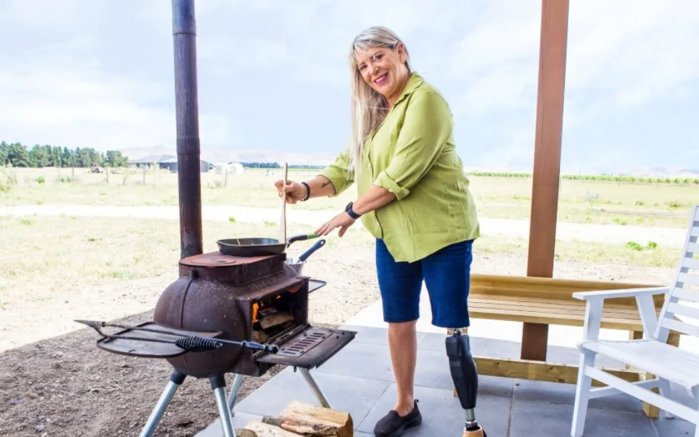 Woman with prosthetic leg cooking outdoors on a wood-burning stove, smiling at the camera.