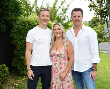 Family of three smiling outdoors, standing on grass with trees in background.