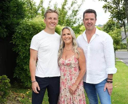 Family of three smiling outdoors, standing on grass with trees in background.