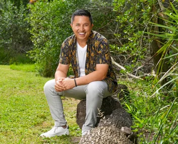 Person smiling, sitting on a log in a green, leafy outdoor setting.