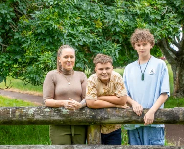A woman and two boys smile while leaning on a wooden fence with lush green trees in the background.