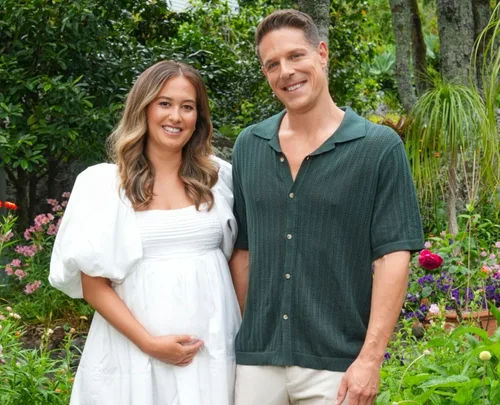A smiling couple poses in a garden, with the woman in a white dress holding her belly.