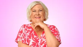 A woman with short blonde hair, smiling in a pink floral top against a pink background.