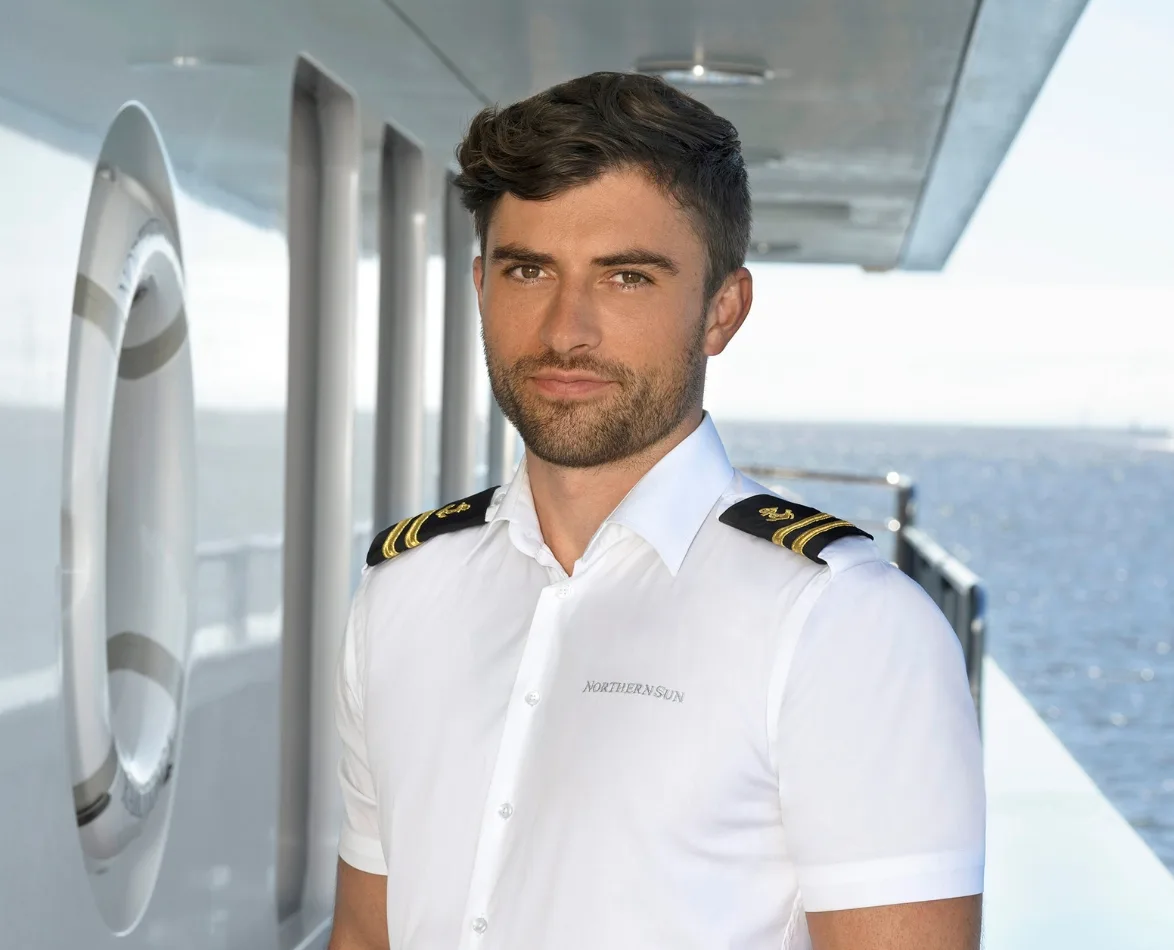 Young man in a white officer's uniform stands on a yacht deck with the ocean in the background.