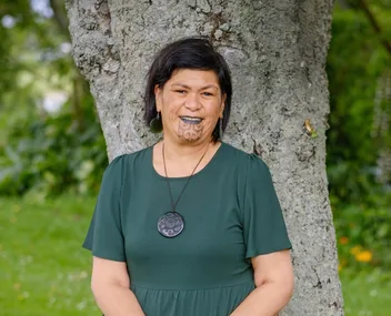 Person with traditional chin tattoo, wearing a green dress and a circular pendant, standing against a tree outdoors.