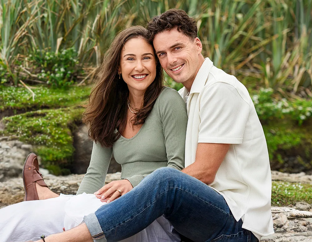 A couple sits outdoors on a stone surface, smiling at the camera, with greenery in the background.
