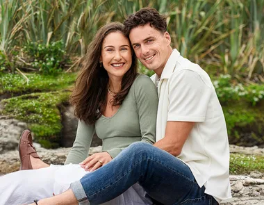 A couple sits outdoors on a stone surface, smiling at the camera, with greenery in the background.