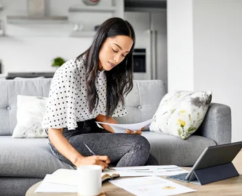 Woman working from home on couch with papers and tablet.