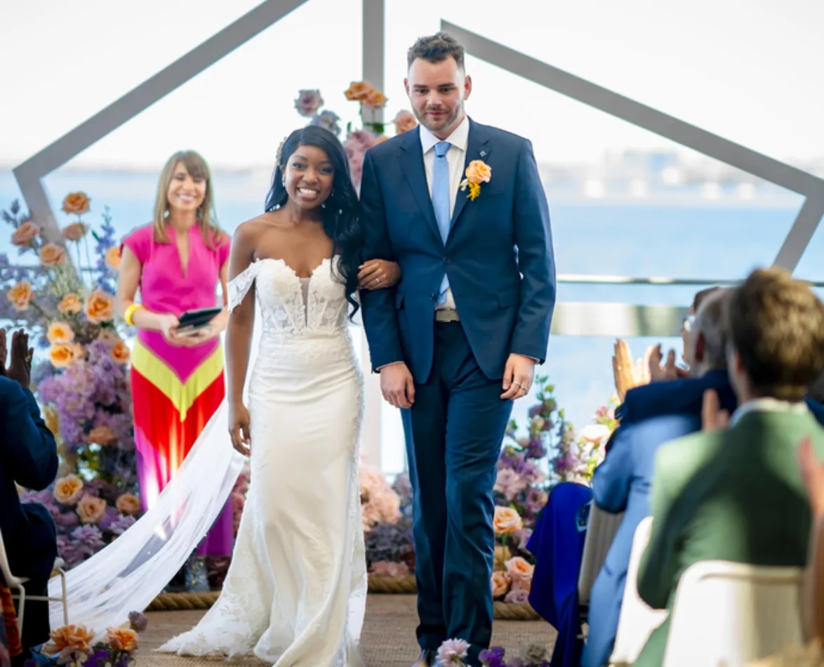 A newlywed couple walks down the aisle, with flowers and a seated audience in the background.