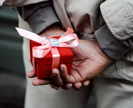A person holding a small red gift box with a pink ribbon behind their back.