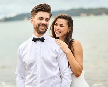 A couple smiling by the water, with the woman resting her hand on the man's shoulder.
