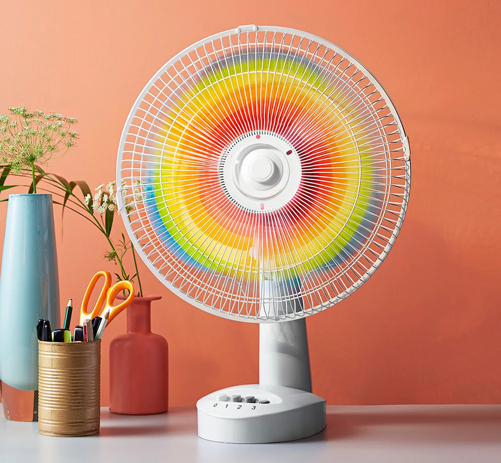 A colorful table fan with rainbow blades on a desk with vases, scissors, and plants against an orange wall.
