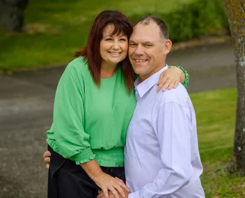 A smiling couple posing outdoors, with greenery in the background. The woman wears a green blouse, and the man a white shirt.