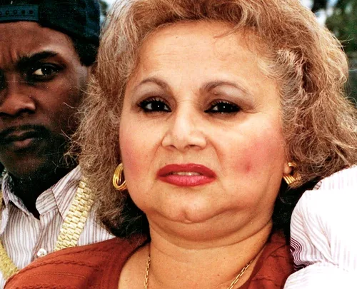 A woman with curly hair and gold earrings looks at the camera; a man wearing a cap is in the background.