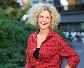 Smiling person with curly hair in red plaid shirt, outdoor setting with greenery in background.