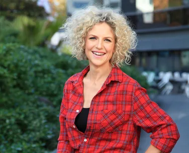 Smiling person with curly hair in red plaid shirt, outdoor setting with greenery in background.