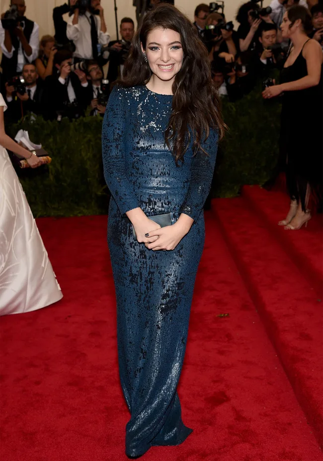 Young woman in a sparkling blue gown smiling on a red carpet surrounded by photographers.