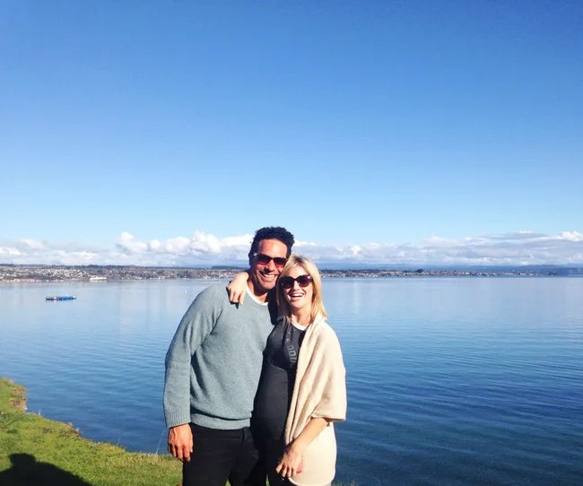A smiling couple in sunglasses stands by a calm lake under a clear blue sky.