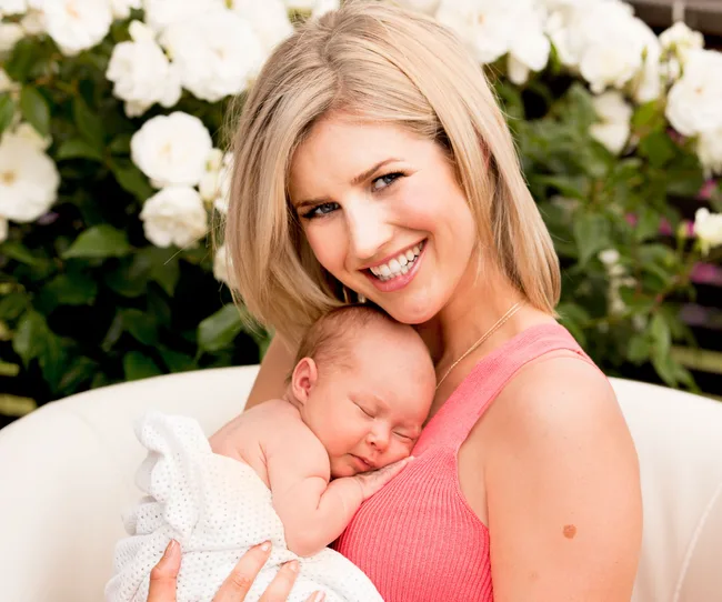 A smiling woman holding a sleeping newborn wrapped in a white blanket, with white flowers in the background.