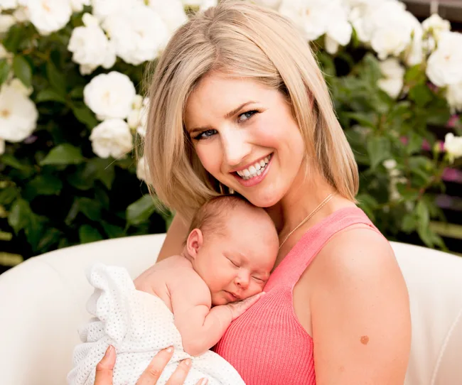 A smiling woman holding a sleeping newborn wrapped in a white blanket, with white flowers in the background.