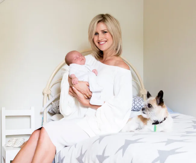 A woman in a white dress sits on a bed holding a sleeping baby, with a small dog beside her.