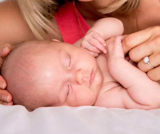Sleeping baby held by a woman with a diamond ring, lying on a soft white surface.