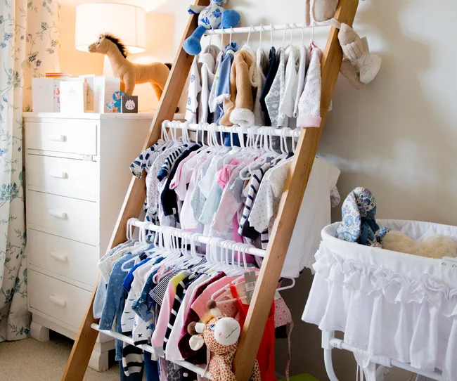 Baby nursery with a wooden rack holding clothes, plush toys on a dresser, and a white bassinet with stuffed animals.