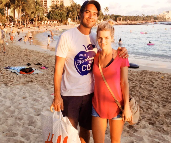 Two people posing on a busy beach, with others swimming and walking in the background.