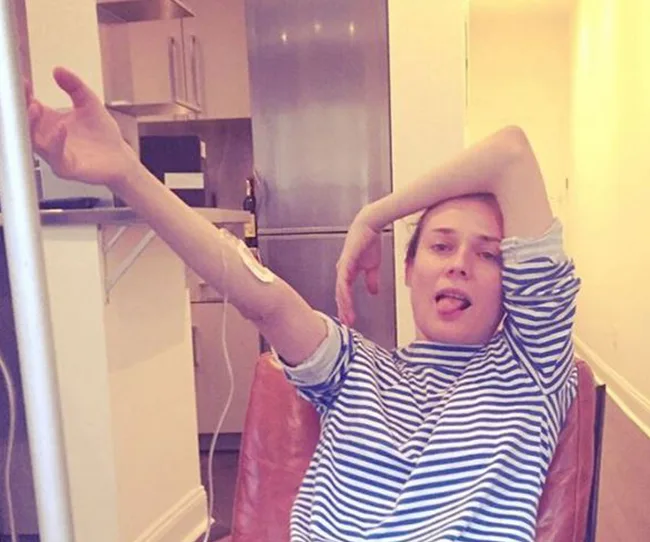 Person in a striped shirt lounging in a chair, arm raised with a medical patch on arm, and tongue slightly out.