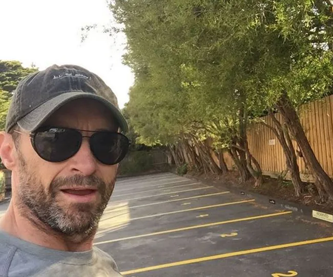 Man in sunglasses and cap takes a selfie in an empty parking lot with trees and a wooden fence in the background.