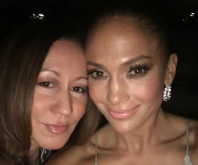 Two women taking a selfie, one smiling with sparkling earrings, both with styled hair and subtle makeup.