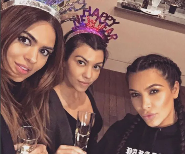 Three women wearing "Happy New Year" headbands, holding champagne glasses, and posing for a selfie indoors.