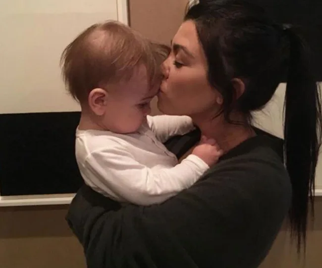 A woman with dark hair kisses a baby on the forehead in a tender moment indoors.
