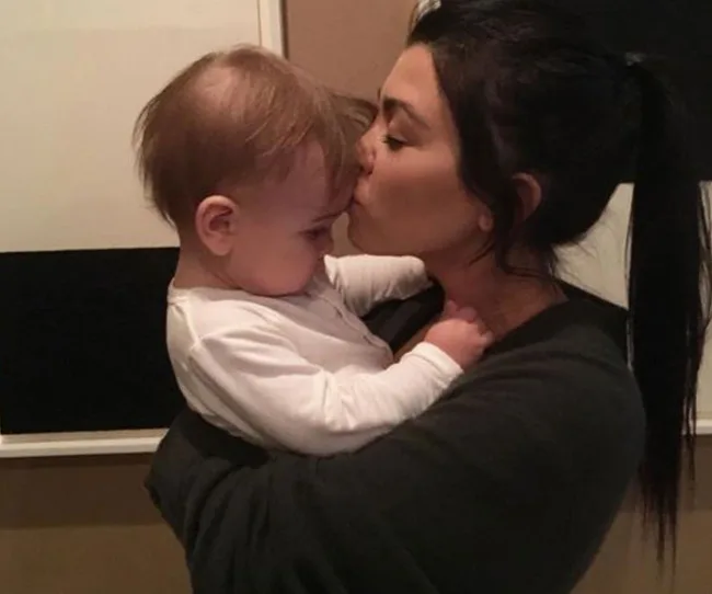 A woman with dark hair kisses a baby on the forehead in a tender moment indoors.