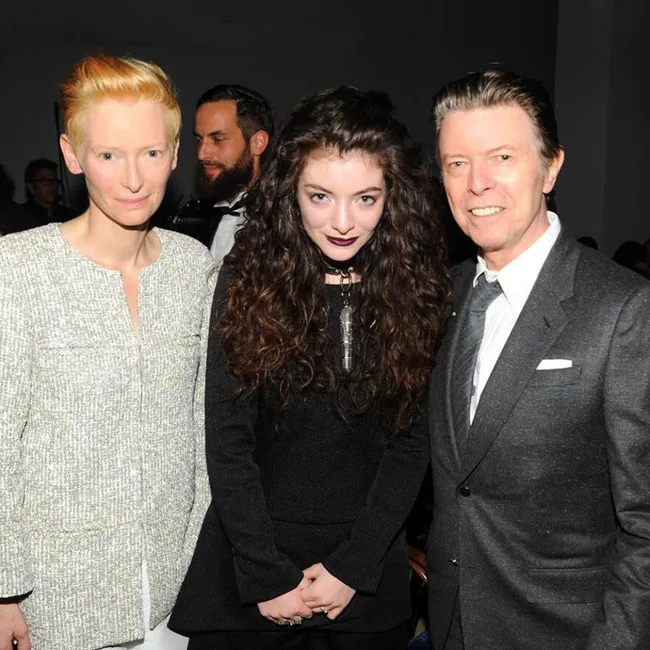 Three people posing closely together at an indoor event. The middle person has long curly hair.
