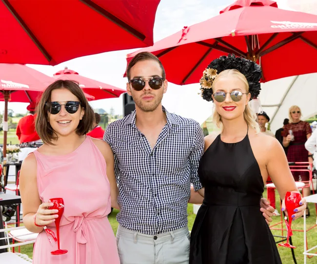 Three people in sunglasses smile for a photo under red umbrellas at an outdoor event, holding red drinks.