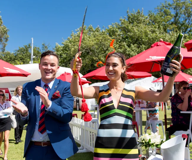 Two people in festive attire celebrating outdoors, woman holding a sword and champagne bottle, surrounded by red umbrellas.