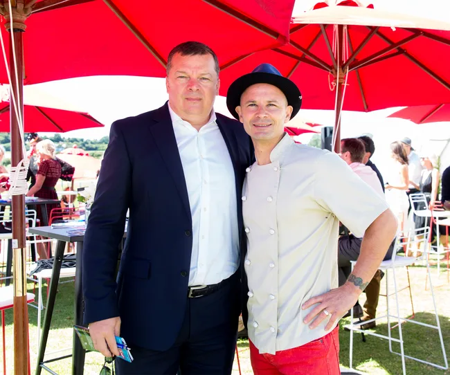 Two men posing together outdoors under red umbrellas; one in a suit, the other in a chef's uniform and hat.