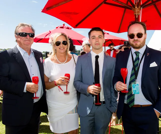 Four people in formal attire holding red glasses, standing under red umbrellas on a sunny day.