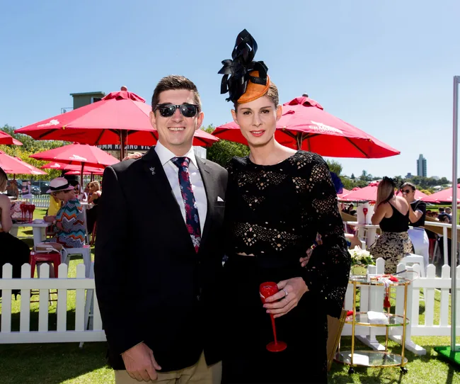 Man in a suit and woman in a black dress and hat smiling outdoors at an event with red umbrellas.