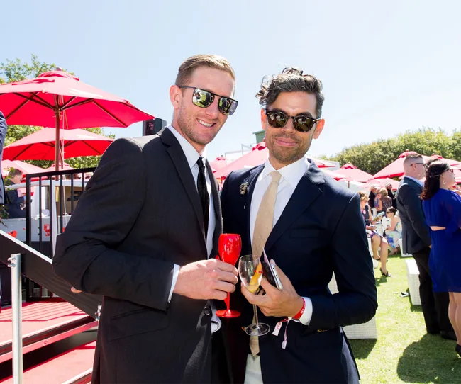 Two men in suits, holding drinks, smiling at a sunny outdoor event with red umbrellas in the background.