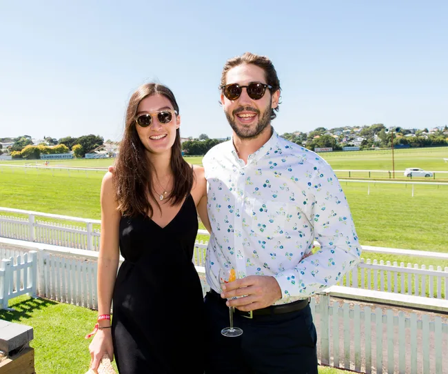 Young couple smiling outdoors at a sunny racecourse, both wearing sunglasses.