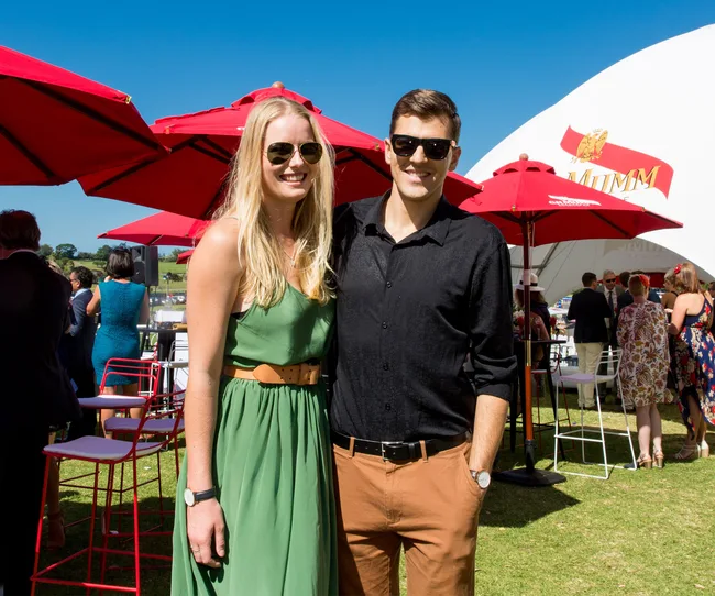 Man and woman smiling outdoors at an event with red umbrellas and a Mumm tent in the background.