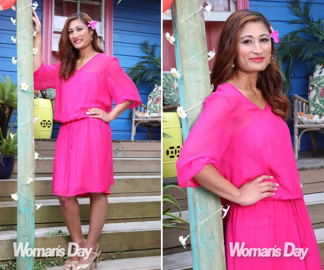 Woman in a bright pink dress with a flower in her hair, smiling, standing on wooden steps with a colorful background.